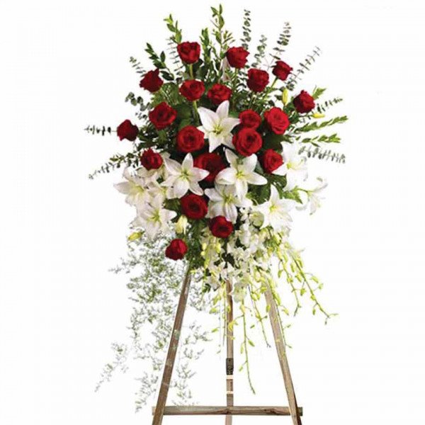 Standing funeral wreath with red roses, white lilies, and white orchids on a wooden easel.