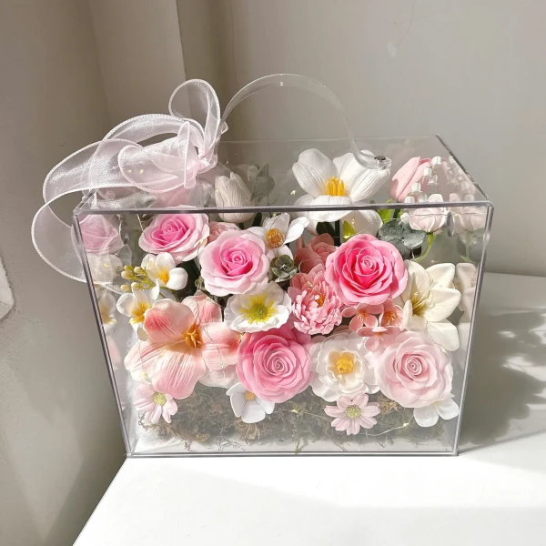 Clear box filled with pink and white artificial flowers, ribbon, and fairy lights.