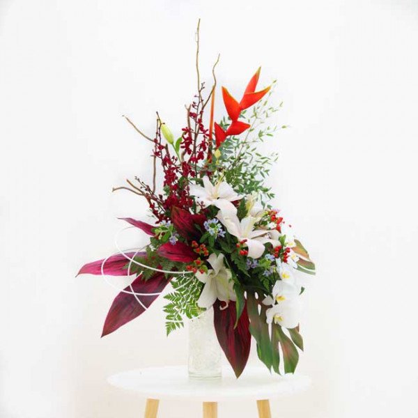 Tropical floral arrangement with lilies, orchids, heliconia, and foliage in a vase.