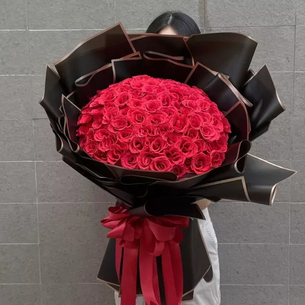A large bouquet of 99 red roses wrapped in black paper with a red bow.