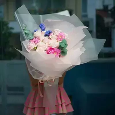 A bouquet of pink and white roses with blue hyacinth, wrapped in white paper, held by a person in a pink dress.