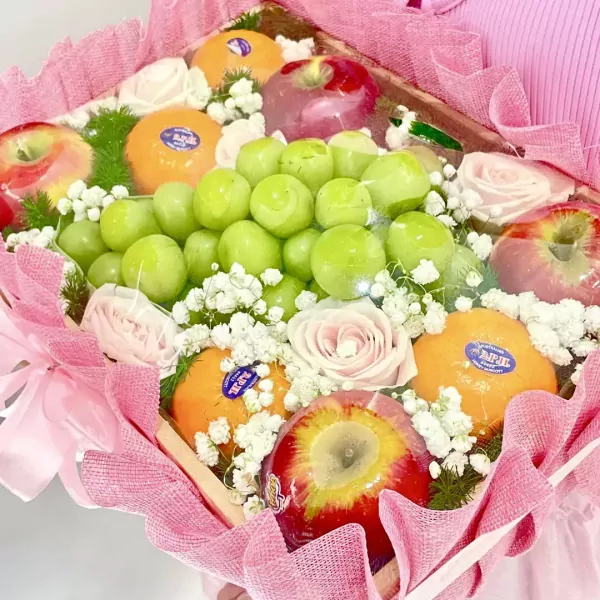 Fruit basket with green grapes, oranges, red apples, and pink roses, wrapped in pink mesh.