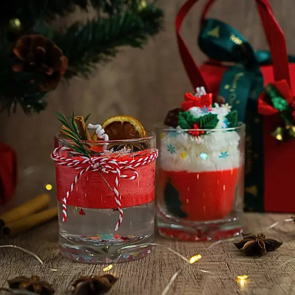 Two Christmas-themed candles in glasses, one with cinnamon stick and dried orange slice, the other topped with whipped cream and festive decorations.