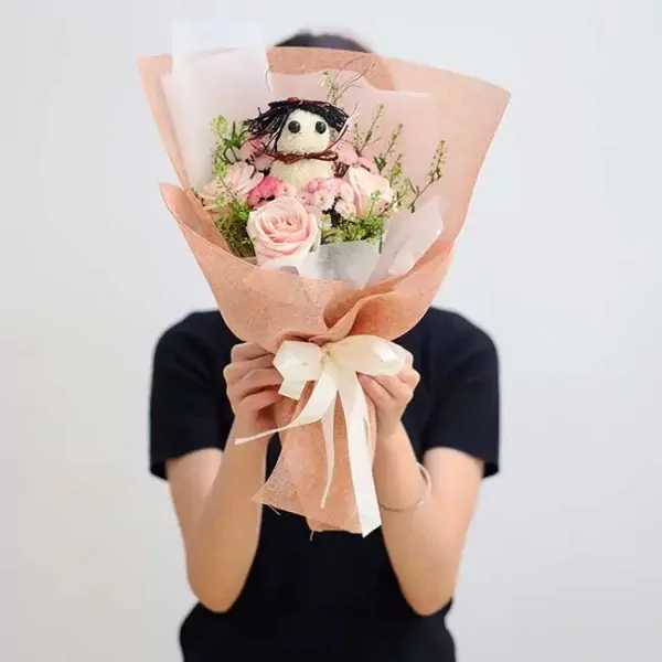 Bouquet with a flower doll on top, pink roses and greenery, wrapped in peach paper, tied with a white ribbon.