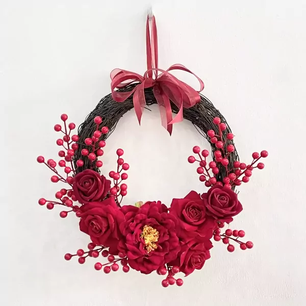 Red rose and berry wreath with ribbon bow hanging on a white wall.