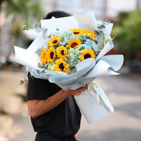 Sunflowers and hydrangeas in a light blue bouquet, held by a person in a black shirt.