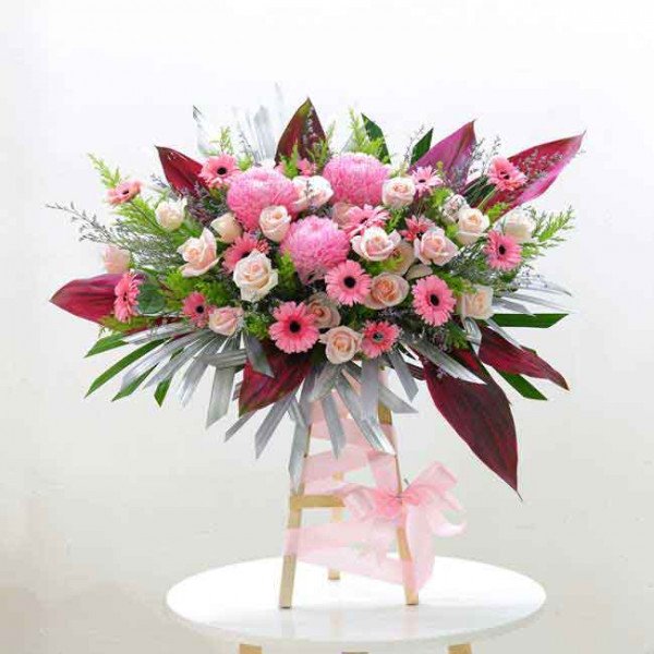 A large, elaborate floral arrangement with pink and cream roses, chrysanthemums, and gerbera daisies on a wooden stand.