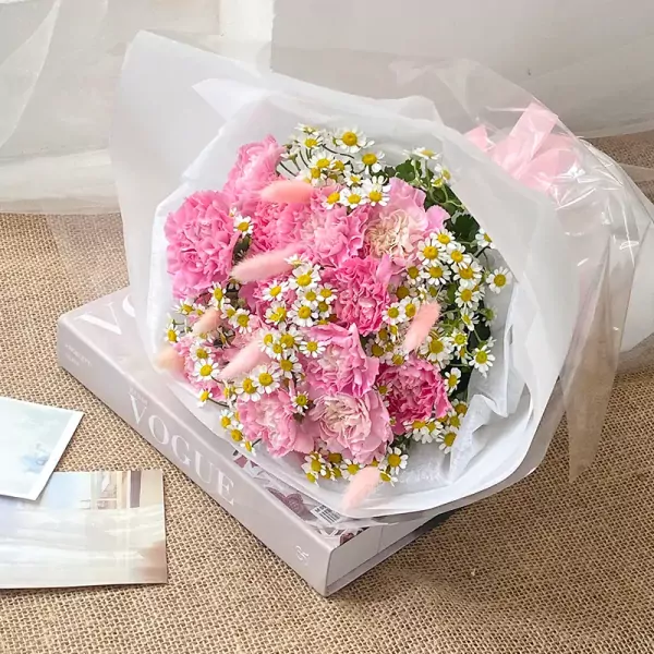 Pink carnation bouquet with daisies and bunny tails, wrapped in white and clear paper, resting on a Vogue book.