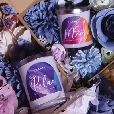 Two scented candles, one labeled "Thank Mom" and the other "Time to Relax," nestled among blue and purple flowers.