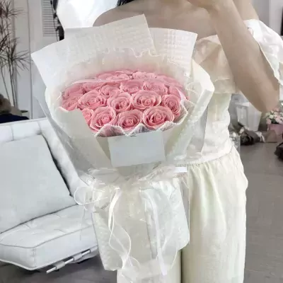 A woman in a light pink dress holds a glamorous pink rose bouquet wrapped in sheer paper and ribbon.