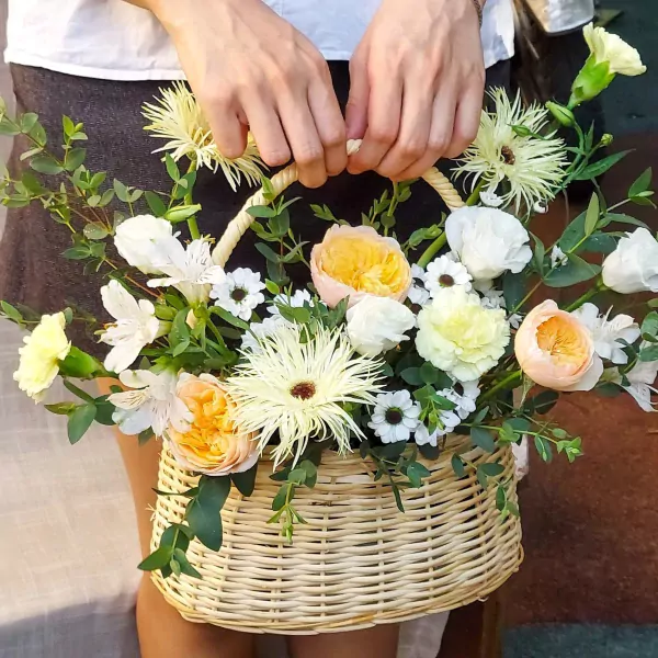 Wicker basket of mixed flowers in peach, yellow, and white, held by hands.